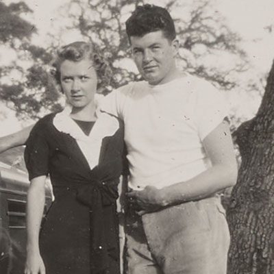 A black and white photograph of a man and a woman dressed casually standing together by a tree.