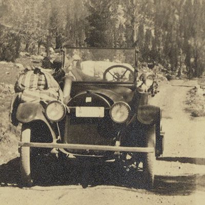 A black and white photograph of an older-style car on a wooded dirt road.