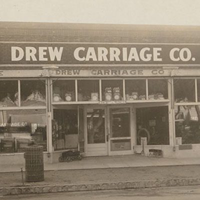 A black and white photograph of a storefront that reads "Drew Carriage Co."