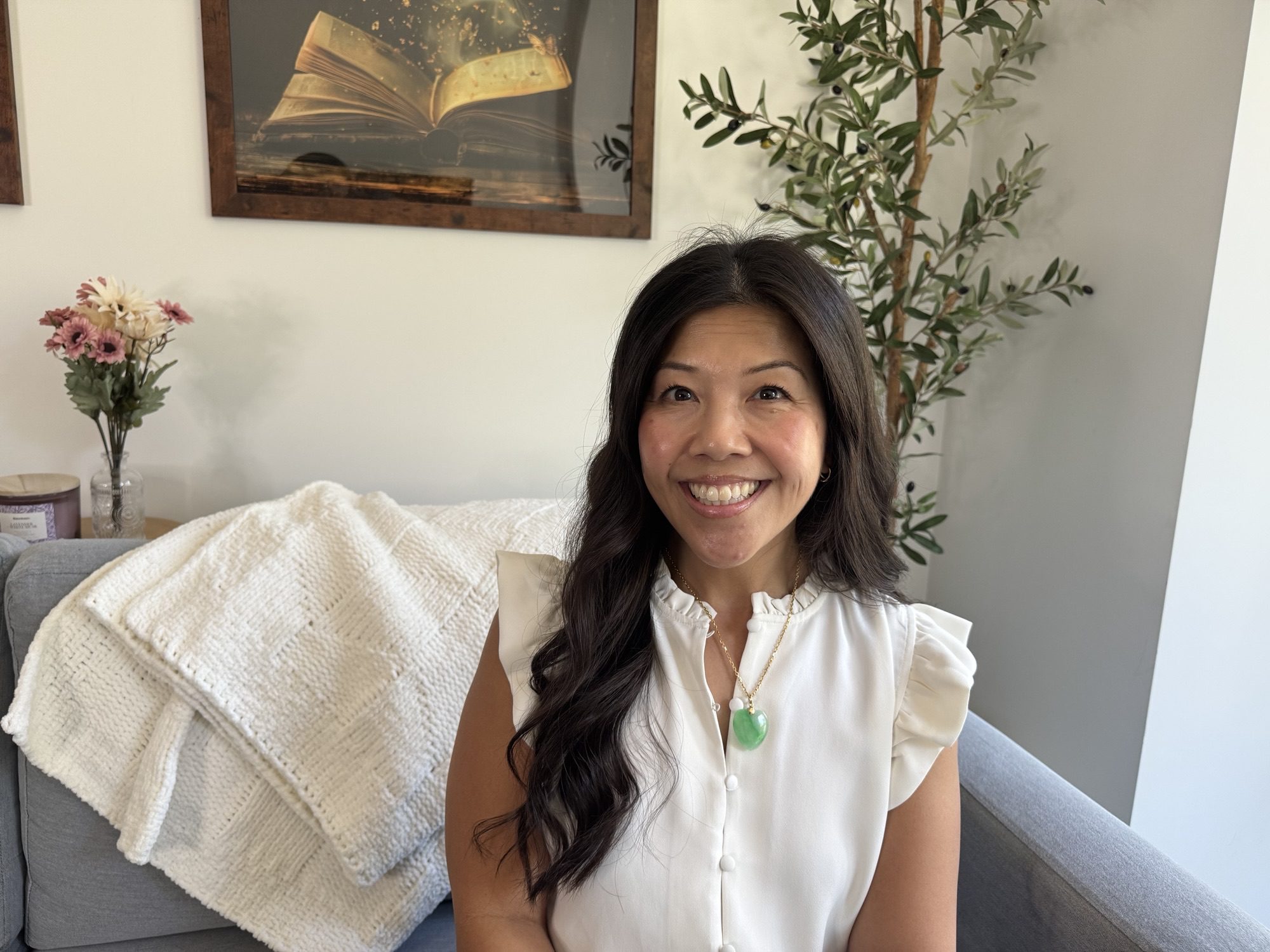 Tammy Myers seated on a sofa with a blanket draped on it, with a framed portrait of an open book, a vase and a tree in the background.
