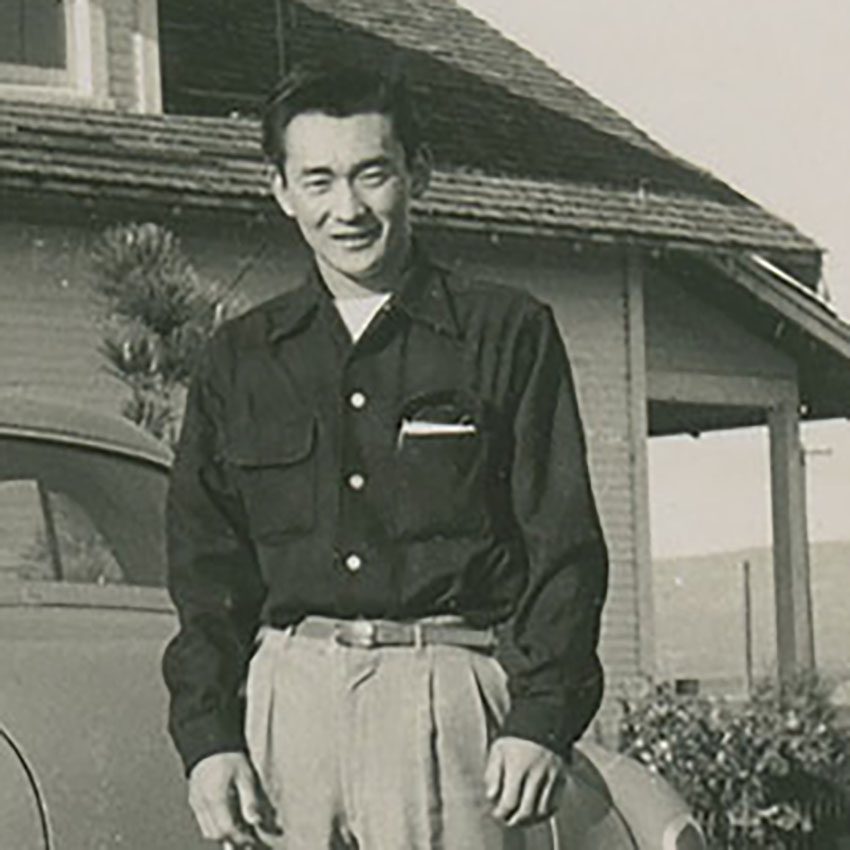 A black and white photograph of a young man standing outside a home beside a car.