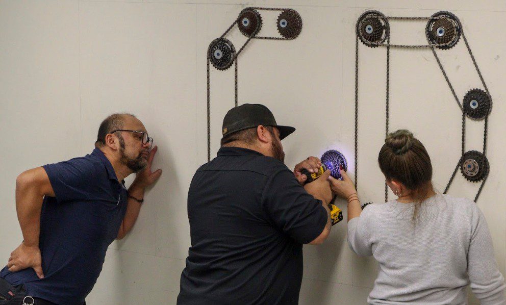 Three people work together to mount a bike hub on a wall, using an electric drill, to create the letter "C" from bike chains and hubs.