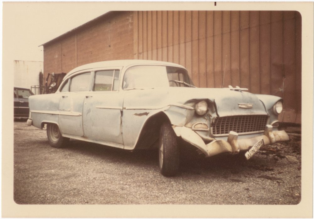 A faded color photograph of a 1950s car with a dented bumper and front headlight