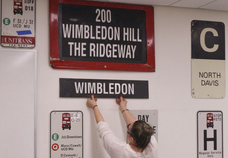 A woman holds up a black bus sign with white lettering that reads "Wimbledon" in front of a wall filled with other bus stop and bus signs.
