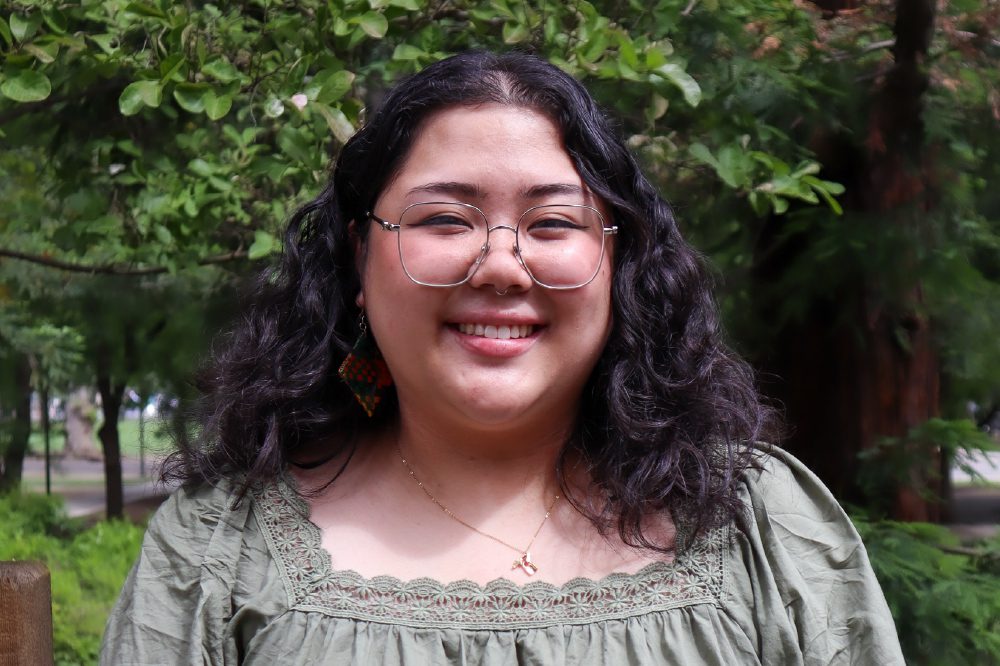 Rianna Herrera poses wearing glasses and a gray-green blouse, with green trees in the background